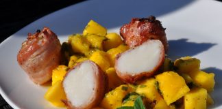 Drei Jakobsmuscheln im Speckmantel neben gelbem Mango-Minz-Chutney mit frischen Minzblättern auf weißem Teller
