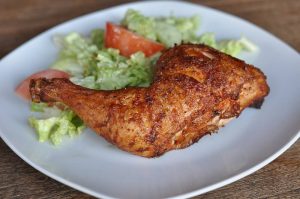 Gegrillter Hähnchenschenkel mit knuspriger, braun gebräunter Oberfläche auf weißem Teller mit grünem Salat und Tomatenscheibe