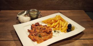 Gyros überbacken in Metaxa-Sauce auf weißem Teller mit Pommes frites, Oliven, Tzatziki und Kapern. Im Hintergrund Knoblauch und Gewürzmischung.
