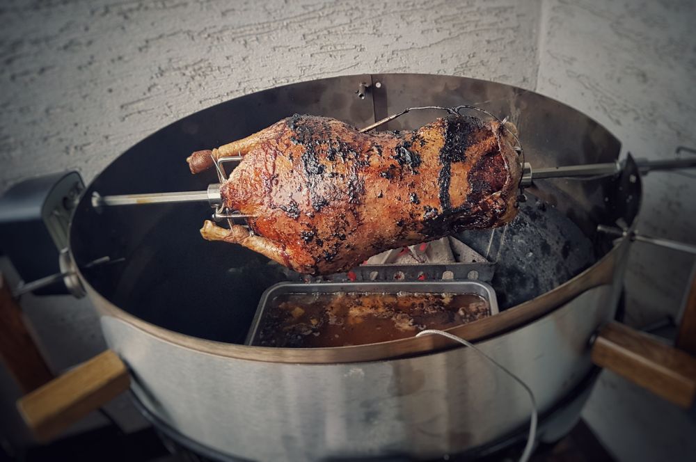 Gegrillte Weihnachtsgans am Drehspieß in Kugelgrill, knusprig gebräunt mit Räucherspuren, Tropfschale darunter