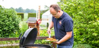 Mann grillt Gemüse und Fleisch auf Kugelgrill im Garten, trägt Teller mit gegrilltem Gemüse in der Hand