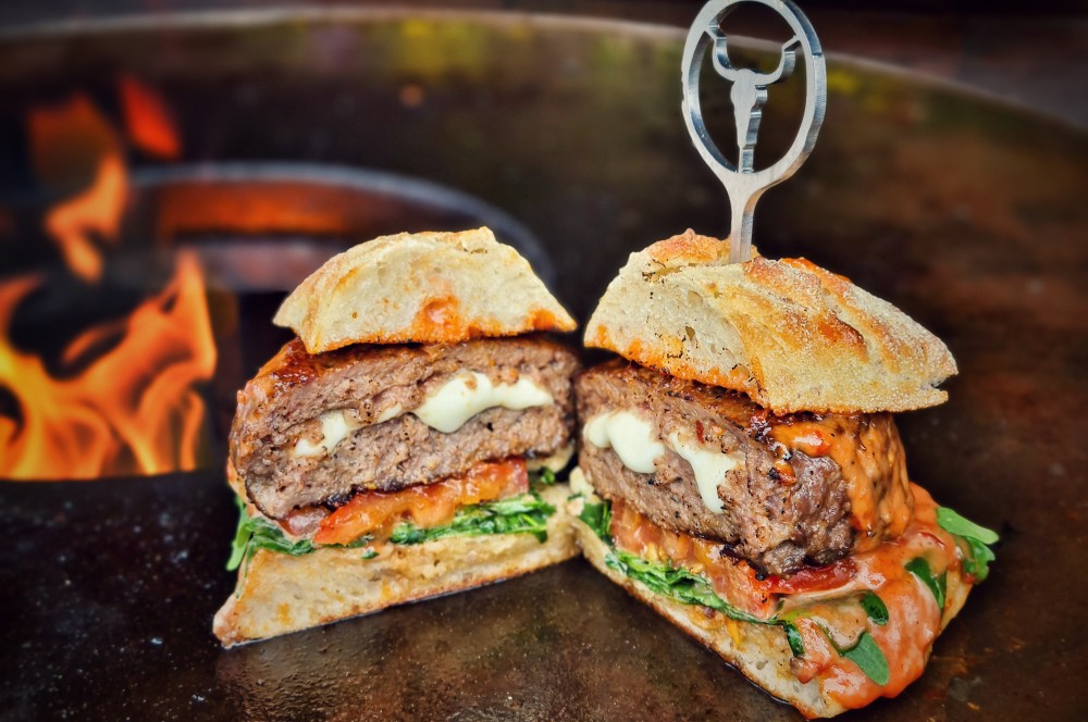 Zwei Italienische Burger mit gegrillten Rindpatties, geschmolzenem Mozzarella, gegrillten Tomaten und Rucola auf getoastetem Burgerbrötchen, gehaltert mit Grillmarks, vor offener Flamme