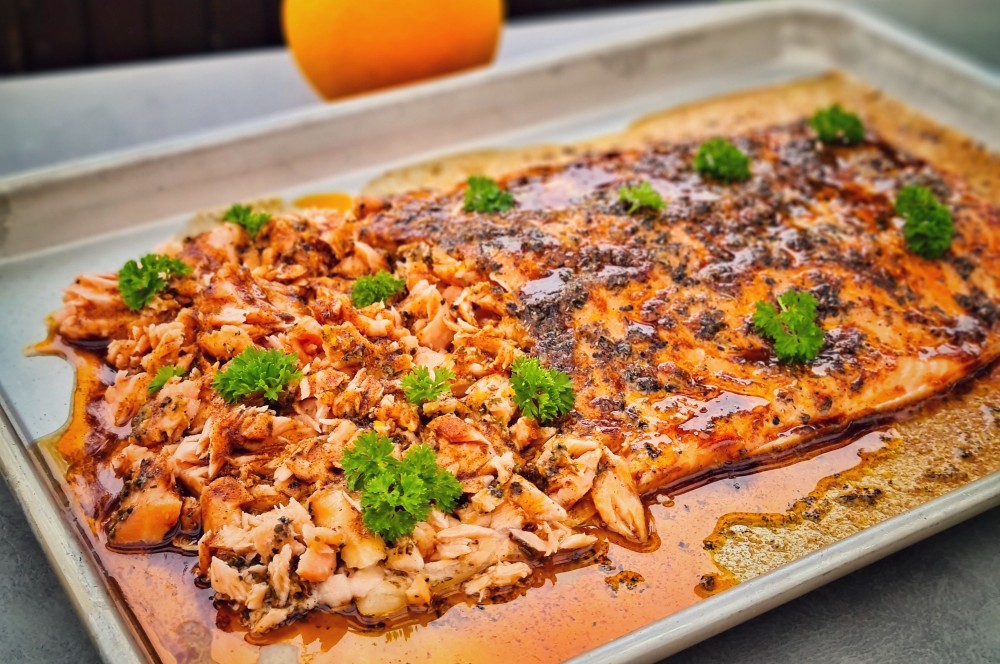 Gegrillter Lachs mit Pulled-Salmon-Topping auf weißem Tablett, garniert mit Petersilie und Pimentón-Öl, Orange im Hintergrund