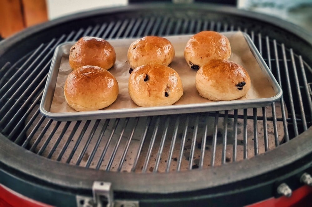 Sechs goldbraune Rosinenbrötchen auf Backpapier in Grillschale auf Kugelgrill, mit schwarzen Rosinen