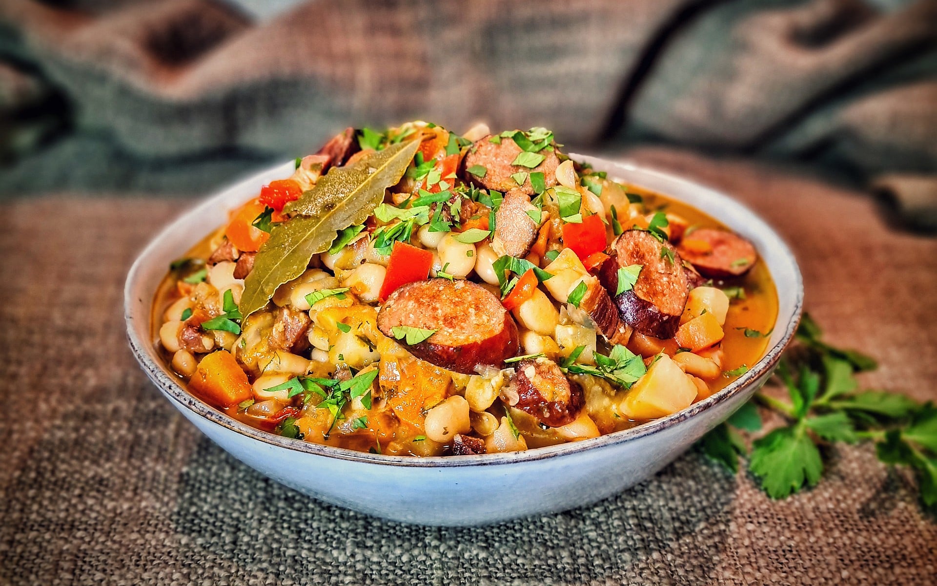 Serbische Bohnensuppe mit Wurst, Kartoffeln, Mais und roten Paprika in weißer Schüssel, garniert mit Petersilie und Lorbeerblatt