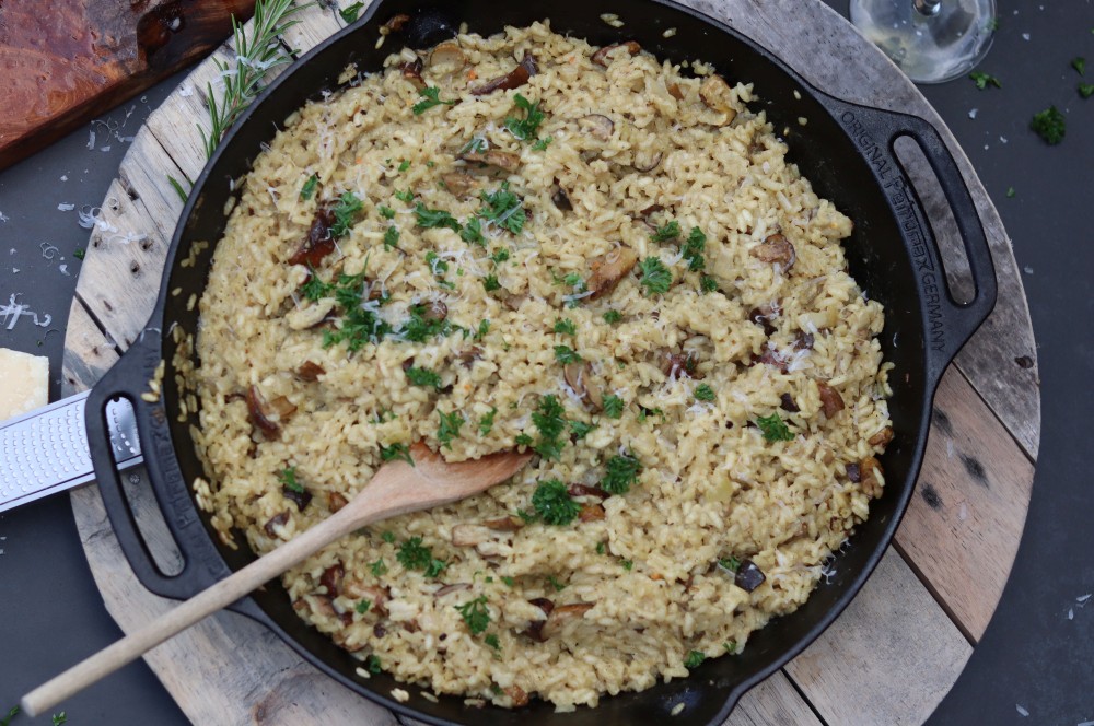 Waldpilz-Risotto in schwarzem Gusseisen-Topf auf Holzuntergrund, cremig mit Waldpilzen und frischer Petersilie garniert, Holzkochlöffel zum Rühren