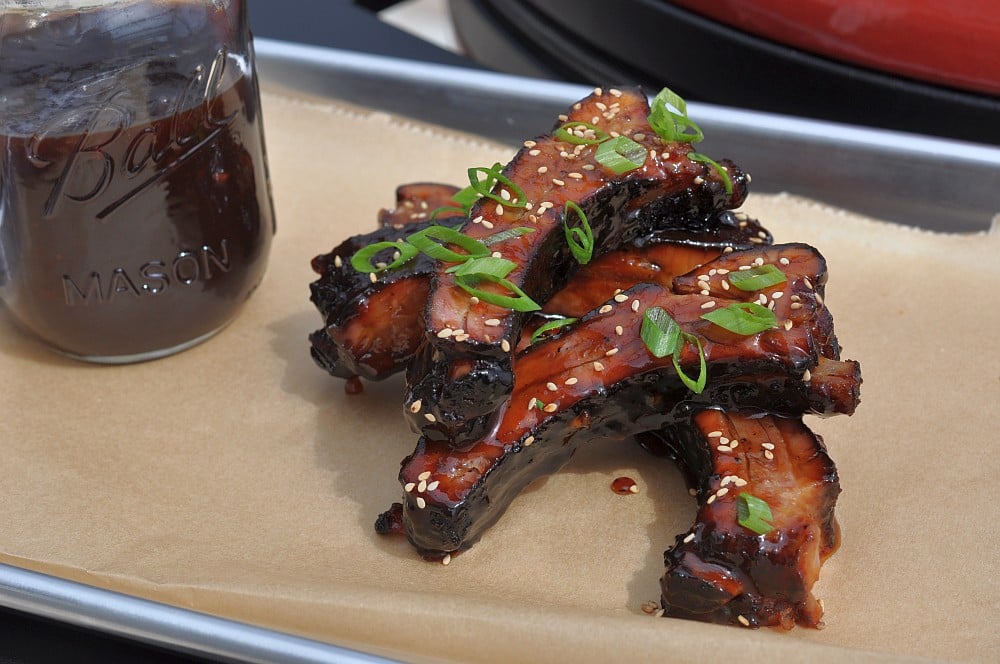 Gegrillte Asian-Sticky-Ribs mit glänzender, dunkler Glasur, Sesam und Frühlingszwiebeln auf Schneidbrett, daneben Glas mit dunkler Sauce