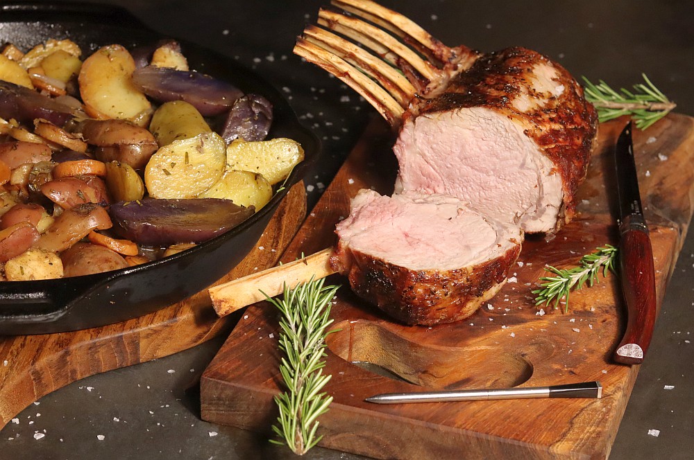 Gegrilltes Kalbs-Karree, aufgeschnitten auf Holzbrett, rosa Fleischkern sichtbar. Daneben Gusseisen-Pfanne mit gegrilltem Wurzelgemüse: Kartoffeln, Karotten und Aubergine. Rosmarin und Fleischbesteck als Deko.