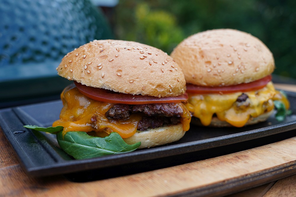 Zwei Double Smashburger mit Sesambrötchen, je zwei dünne Beef-Patties, geschmolzenem Cheddar, Tomatenscheibe und Rucola auf schwarzem Servierbrett
