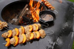 Hähnchen-Pfirsich-Spieße auf schwarzer Feuerplatte grillen, daneben kleine Gusseisenpfanne mit Glasur, offenes Feuer im Hintergrund