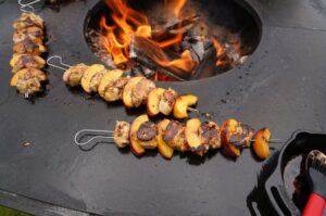 Hähnchen-Pfirsich-Spieße auf schwarzer Grillfläche über offenem Feuer, Fleisch und Pfirsichstücke mit Röstfarbe