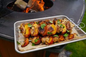 Gegrillte Hähnchen-Pfirsich-Spieße mit Paprika auf weißem Tablett, garniert mit grünen Kräutern, im Hintergrund Feuer in der Feuerschale