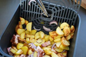 Bauernfrühstück in Heißluftfritteuse: Kartoffeln und Speckstücke in schwarzem Frittierkorb, oben Grillrost mit roten Zwiebeln und Bratelementen