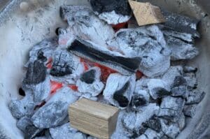 Glühende Holzglut mit Kohle und Asche in einem Grill, daneben frisches Holzscheit für Räuchertemperatur