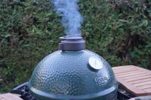 Kamado Joe Keramikgrill mit geschlossenem Deckel und Rauch, auf Holztisch, im Hintergrund grüne Hecke