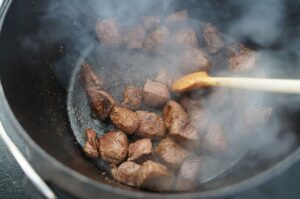 Rindergulaschstücke im Dutch Oven werden mit Holzlöffel gerührt, Fleisch gart im Rauch