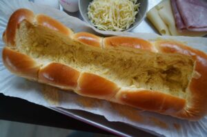 Gefüllter Brioche-Hefezopf mit Ei und Käse, daneben geriebener Käse, Schinken und Käsescheiben auf Backpapier