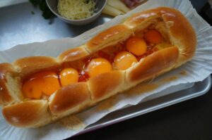 Gefüllter Brioche-Hefezopf mit Spiegeleiern und geschmolzenem Käse, daneben geriebener Käse und Schinken