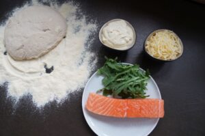 Zutaten für Lachs-Pizza: Pizzateig, Creme fraîche, geriebener Käse, frischer Lachs und Rucola auf dunkler Arbeitsfläche