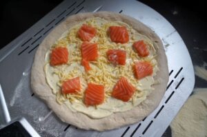 Lachs-Pizza mit Käse und Lachsfilet-Stücken auf Pizzastein vor dem Grillen