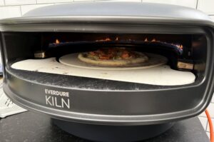 Everdure Kiln Pizzaofen mit brennendem Feuer, auf dem Pizzastein eine fertig gebackene Lachs-Pizza mit grünem Belag, daneben weißer Pizzastein