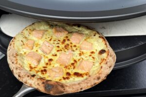 Lachs-Pizza mit Käse und Lachsstücken auf schwarzem Grillstein im Smoker, knusprig gebräunter Teigrand