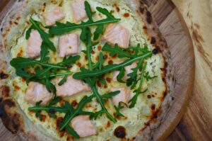 Lachs-Pizza mit Rucola auf Holzbrett. Käsiger Belag mit rohen Lachsstücken und frischen Rucola-Blättern. Knusprig gebräunte Kruste.