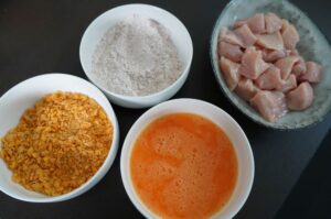 Zutaten für Chicken Nuggets: gewürfelte Hähnchenbrust, Cornflakes-Panade, Mehl und verquirltes Ei in weißen Schüsseln