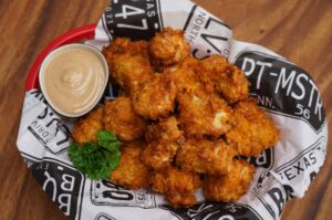 Knusprige Chicken Nuggets mit Cornflakes-Panade auf Papier mit BBQ-Pit-Logo, garniert mit Petersilie und Dip in roter Schale