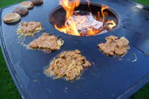 Smashburger-Patties mit karamellisierten Zwiebeln auf schwarzer Grillplatte über offener Flamme, daneben Burger-Brötchen zum Toasten