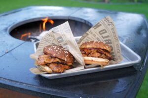 Zwei Smashburger mit Sesambrötchen in Papiertüten auf weißem Tablett vor loderndem Feuer in Feuerschale