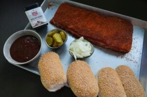 McRib-Zutaten auf weißem Tablett: gewürzter Rippenbratling mit BBQ PIT Pork Rub, BBQ-Sauce in Schüssel, Sesambrötchen, eingelegte Gurken und Zwiebeln
