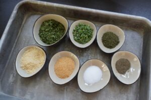 Acht Schalen mit Zutaten für Ranch-Gewürz: getrocknete Kräuter, grüne Zwiebeln, Knoblauchpulver, Zwiebelpulver, Salz und Pfeffer auf Backblech