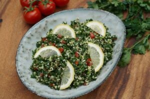 Tabouleh-Salat mit gehackter Petersilie, Bulgur, Tomaten und Zitronenscheiben in grauer Schale auf Holztisch