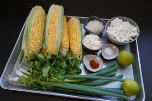 Zutaten für Esquites: vier frische Maiskolben mit Blättern, Koriander, Frühlingszwiebeln, Limonen, Maiskörner, Frischkäse, geriebener Käse, Salz und Paprikapulver auf weißem Tablett.