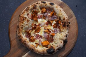 Alpen-Pizza mit geschmolzenem Bergkäse, knusprigem Speck und gebratenen Pfifferlingen auf Holzbrett