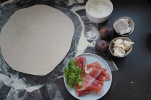 Zutaten für Feigenpizza: ausgerollter Pizzateig, Serrano-Schinkel mit Rucola, Ziegenkäse, rote Feigen und Creme fraîche auf dunklem Untergrund