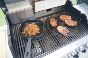 Grill mit Nackensteaks und karamellisierten Miso-Zwiebeln in Grillpfanne, mehrere angebratene Steaks auf Grillrost
