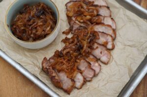 Gegrillte Nackensteaks in Scheiben geschnitten auf Backpapier mit karamellisierten Miso-Zwiebeln in weißer Schale