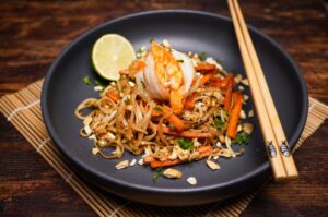 Pad Thai mit gegrillten Garnelen, Reisnudeln, Erdnüssen und frischen Kräutern in schwarzer Schale, garniert mit Limette und Stäbchen