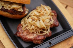 Thüringer Rostbrätel mit knusprigen Zwiebelringen auf schwarzem Teller, im Hintergrund ein Brötchen mit Zwiebelbelag