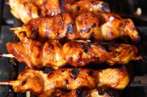 Yakitori-Spieße mit mariniertem Hähnchenfleisch auf dem Grill, goldbraun und mit Grillmarks gebräunt, auf Metallspießen