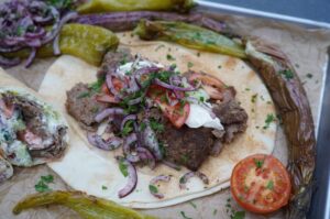 Backpapier-Döner mit gegrilltem Hackfleisch, Joghurt-Sauce, roten Zwiebelringen, Tomaten und Petersilie auf Fladenbrot, umgeben von gegrillten Paprika und Zwiebeln