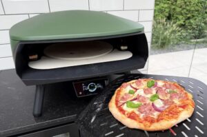 Salsiccia-Pizza mit Mozzarella, Salami und roten Zwiebeln auf Pizzaofen-Kochplatte, grüner Deckel geöffnet.