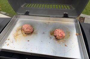 Zwei Rindfleisch-Patties auf Grillplatte, mit Gewürzen bestreut, beim Garen für Béarnaise-Smashburger