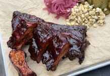 Cadillac-Cut-Spare-Ribs mit dunkler Räucherkruste auf Schneidebrett, garniert mit eingelegten roten Zwiebeln, Essiggurken und Mais