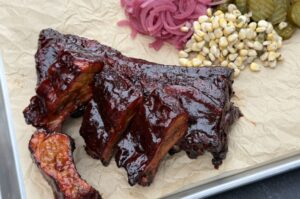 Cadillac-Cut-Spare-Ribs mit dunkler Räucherkruste auf Schneidebrett, garniert mit eingelegten roten Zwiebeln, Essiggurken und Mais