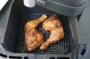 Zwei knusprig braun gebratene Hähnchenschenkel in der Heißluftfritteuse auf schwarzem Gitterrost