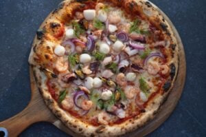 Pizza Frutti di Mare mit Garnelen, Muscheln, roten Zwiebeln, Mozzarella und Dill auf Holzbrett