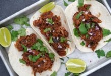 Drei Pulled-Pork-Tacos auf Metallblech mit Koriander, roten Zwiebelringen und Limettenspalten